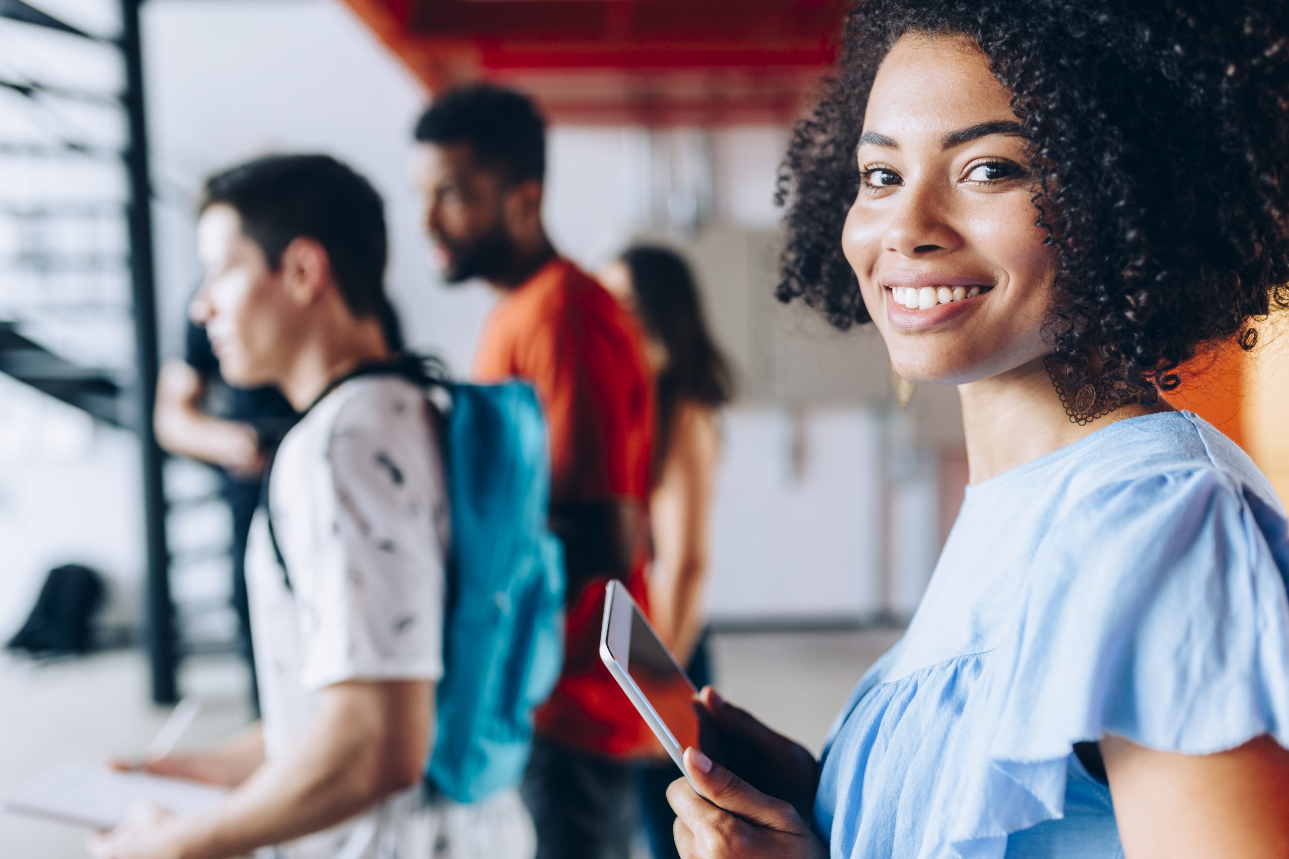 jovens entrando em uma instituição de ensino, com mochilas nas costas. em primeiro plano, uma jovem negra, com cabelos cacheados, sorrindo e segurando um tablet