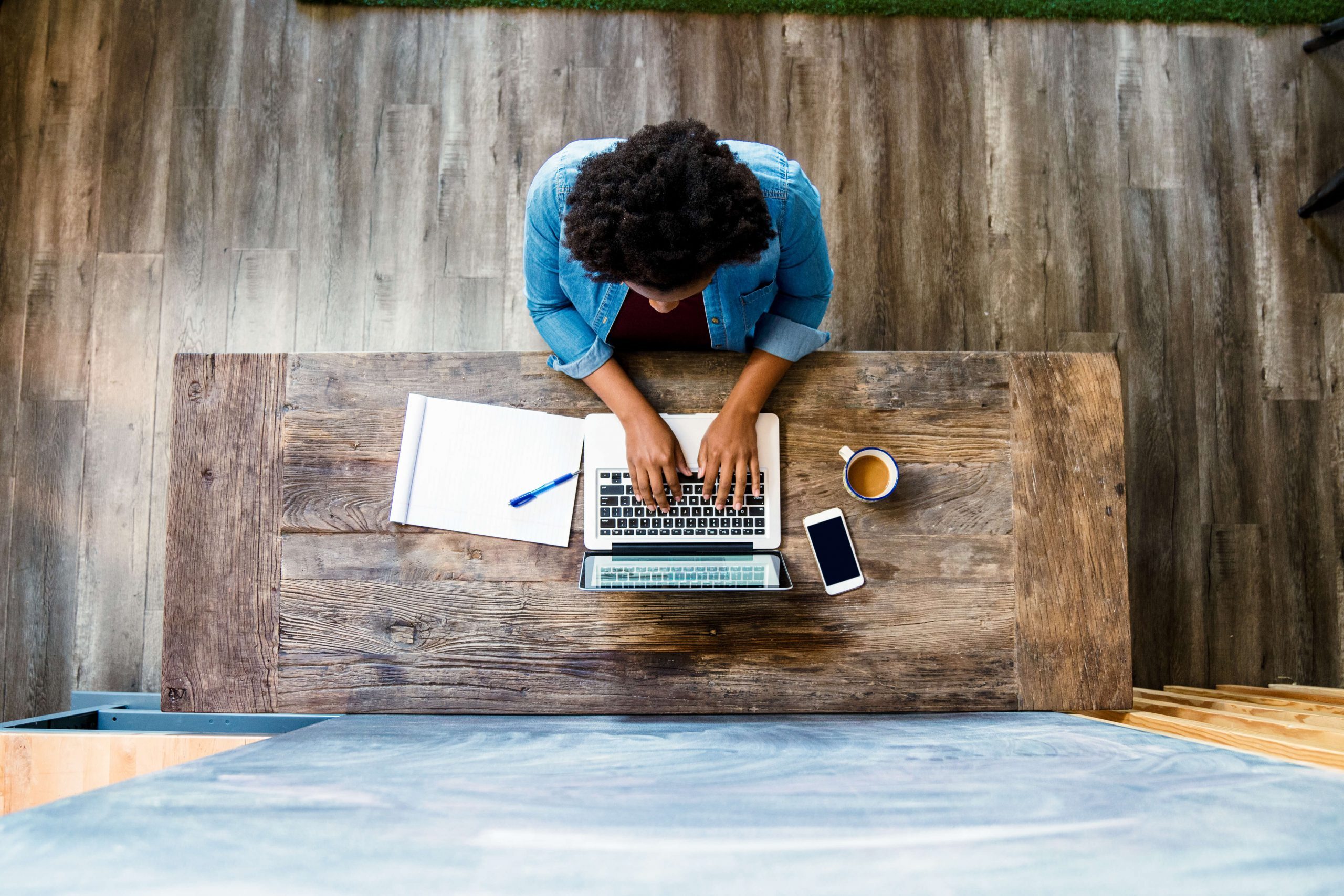 foto tirada de cima, de um profissional em uma mesa de trabalho, trabalhando em um notebook com papel e caneta do lado esquerdo, e do lado direito um smartphone e uma caneca de café