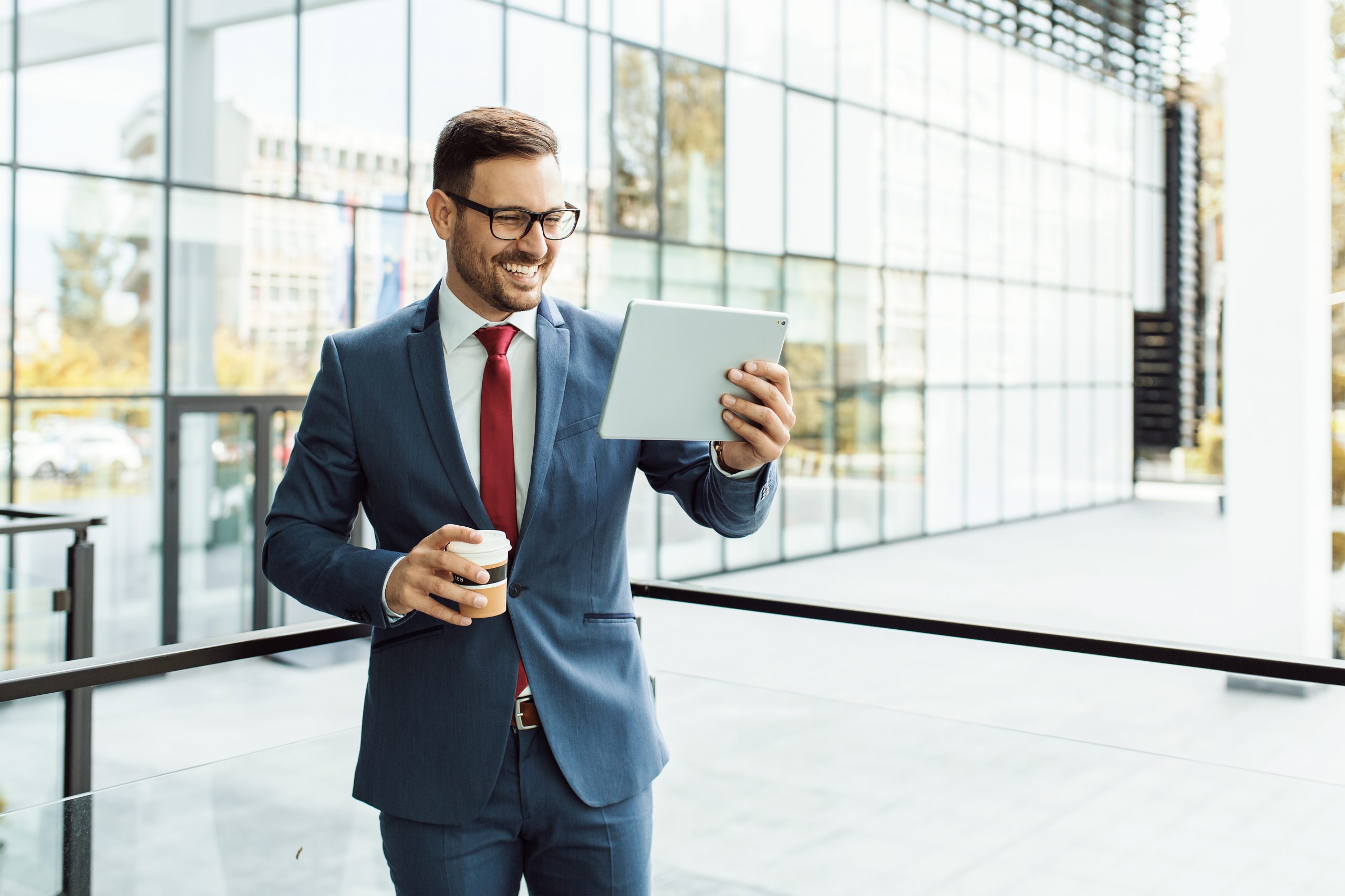 homem branco, com cabelos castanhos e curtos, barba baixa, usando terno azul, gravada vinho e óculos, segurando um café com uma mão e um tablet com a outra, sorrindo enquanto olha para a tela