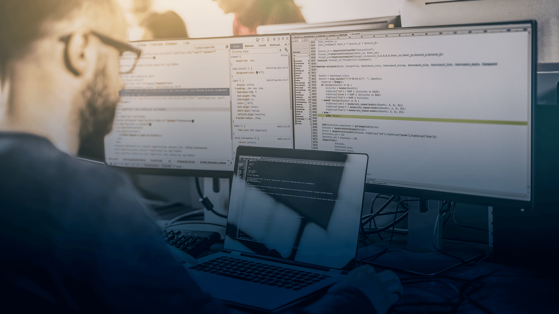 profissional trabalhando em um notebook, com dois monitores exibindo informações atrás do notebook
